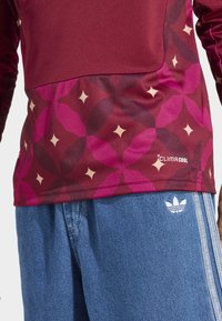 Maroon long-sleeve top with pink geometric pattern and star accents; paired with light blue denim shorts featuring three white stripes and logo.