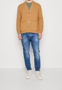 Cárdigan de lana color beige con cuello en V, textura acanalada y dos bolsillos delanteros, combinado con jeans azules y zapatillas claras.