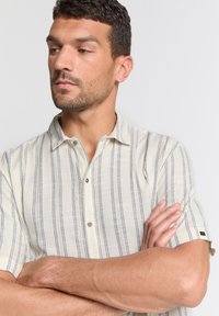 Korte mouwen, colshirt in crème met blauwe verticale strepen. Heeft knopen en een subtiele textuur. Slank fit ontwerp.