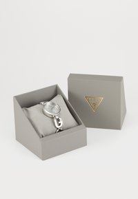 Silver Guess wristwatch with crystal markers on a gray pillow inside a gray box, next to a gray box lid with a gold Guess logo.