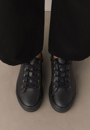 Para czarnych skórzanych sneakersów sznurowanych, noszonych z czarnymi spodniami, widziana z góry na jasnej podłodze.