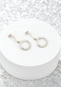 Silverfärgade hoopsörhängen med en polerad cirkulär design och ett texturerat band prytt med klara kristaller. Visas på en vit pedestal.