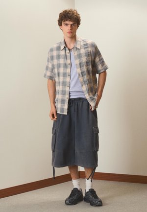 Junger Mann steht drinnen und trägt ein graues kariertes Kurzarmhemd, ein hellgraues T-Shirt, dunkle Cargoshorts, weiße Socken und schwarze Turnschuhe.