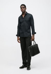 Camicia nera con bottoni abbinata a pantaloni scuri, che tiene una borsa in pelle nera. Design slim fit, tessuto morbido e scarpe lucide.
