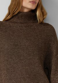 Bruine gebreide trui met een hoge col, voorzien van een gestructureerd ribbelpatroon en een losse pasvorm. Warm en gezellig ontwerp voor casual gebruik.