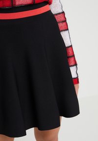 Black skirt with a flared design and a vibrant red elastic waistband, featuring a textured fabric. Minimalist and solid color.