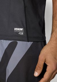 Camisola desportiva preta com etiqueta "Venum". Apresenta tecido que afasta a humidade e costuras planas. Padrão cinzento nas calças curtas realça o design desportivo.