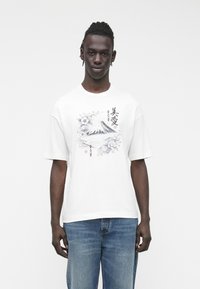 Weißes Baumwoll-T-Shirt mit kurzen Ärmeln, vorne mit einem schwarz-grauen Bergmotiv und floralem Design. Blaue Jeans sichtbar.