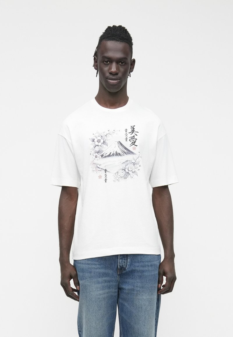 Weißes Baumwoll-T-Shirt mit kurzen Ärmeln, vorne mit einem schwarz-grauen Bergmotiv und floralem Design. Blaue Jeans sichtbar.