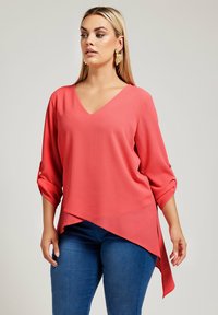 Blusa coral a V con maniche a tre quarti con risvolto, orlo asimmetrico e tessuto leggero, abbinata a jeans blu slim fit.