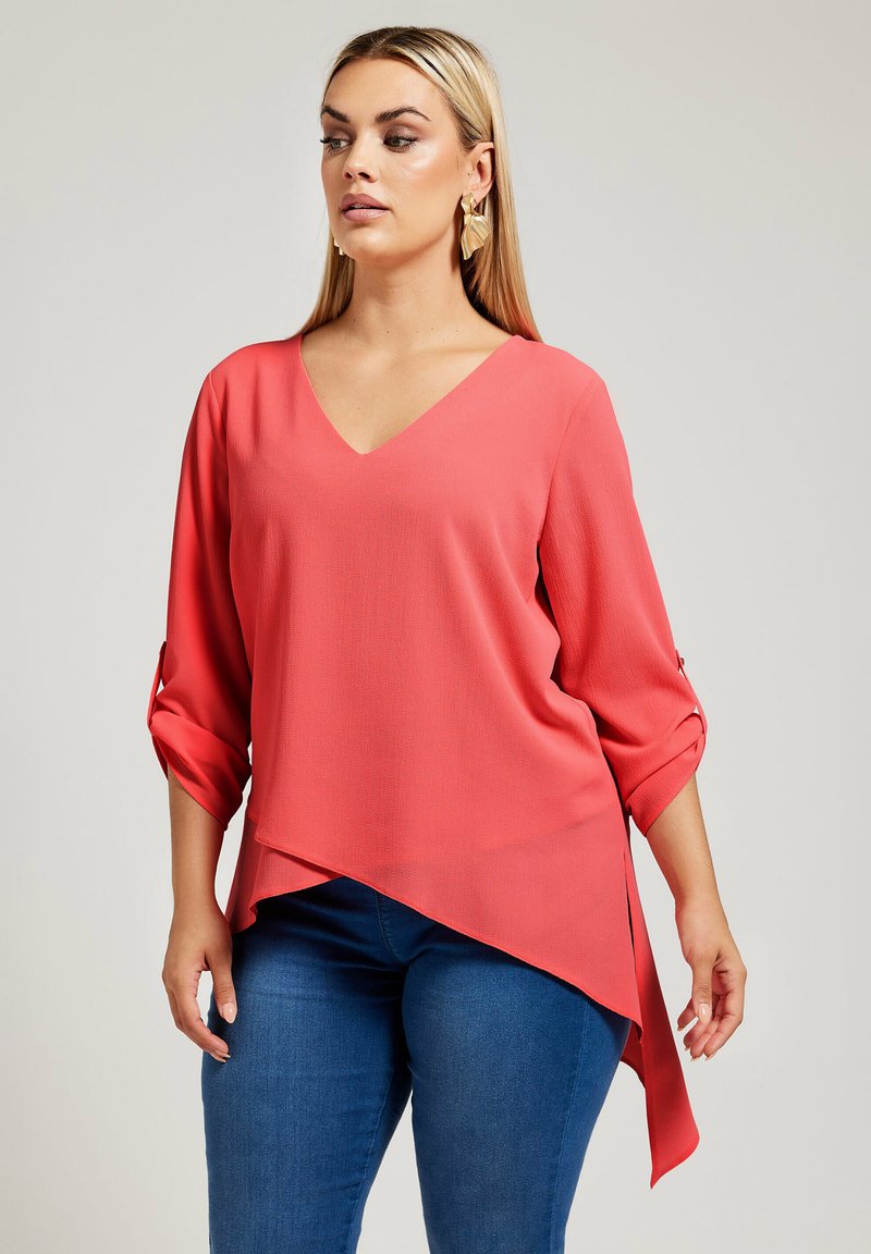 Blusa coral a V con maniche a tre quarti con risvolto, orlo asimmetrico e tessuto leggero, abbinata a jeans blu slim fit.