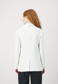 Blazer bianco con un design aderente, spacco singolo sul retro e dettagli di branding discreti sul colletto. Texture liscia, maniche lunghe.