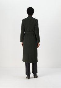 Palton lung din lână verde, cu centură, guler ascuțit și țesătură texturată. Purtați peste pantaloni negri, evidențiind o siluetă ajustată și lungimea până la genunchi.