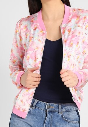 Žena nosí růžovou květinovou bomber bundu přes tmavě modré tričko s výstřihem do V a světle modré džíny, drží bundu otevřenou na zipu.