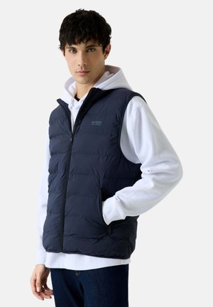 Giovane uomo con capelli scuri che indossa un gilet imbottito blu navy senza maniche sopra una felpa bianca con cappuccio, posando con una mano in tasca.
