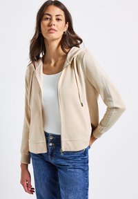 Felpa beige con zip e righe laterali testurizzate, dotata di cappuccio, due tasche e polsini a costine. Abbinata a jeans in denim blu scuro.