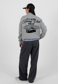 Szara oversize'owa bluza z czarnym napisem i grafiką samochodu na plecach, z prążkowanymi mankietami, noszona na niebieskiej koszuli w paski, zestawiona z czarnymi spodniami.