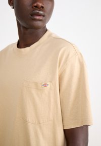 Tricou bej cu mânecă scurtă, fabricat din bumbac, având un buzunar pe piept cu un logo colorat. Textură moale, croială lejeră și decolteu rotund.