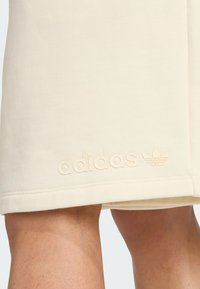 Beżowe szorty sportowe wykonane z miękkiego materiału, z wytłoczonym logo "adidas" oraz symbolem trzech liści marki w okolicy dołu.
