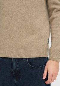 Beige gyapjú pulóver ribbelt mandzsettákkal és alján, sötétkék farmerrel párosítva. A pulóver texturált felülettel és oldalán címkével rendelkezik.