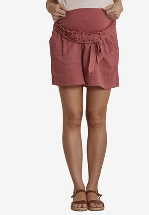 Frau trägt mauvefarbene Umstandsshorts mit gebundenem Gürtel, weißes Oberteil und braune Sandalen, steht vor einem schlichten weißen Hintergrund.