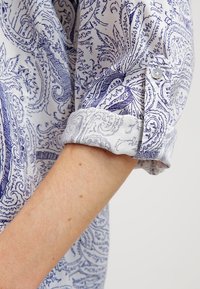 Camicia bianca con un motivo paisley blu, caratterizzata da un risvolto con un accento a bottone. Tessuto liscio con una trama dettagliata.