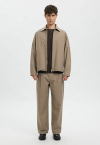 Beige oversized jakke med krave og lynlås, kombineret med matchende bukser med vide ben. Mørk trøje under. Sorte sko fuldender looket.