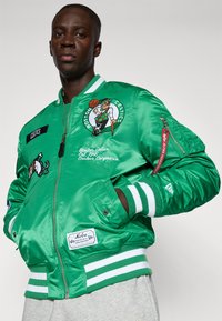 Jachetă bomber din satin verde cu logo-ul Boston Celtics și broderie. Prezintă manșete și talie cu dungi ribbate, două buzunare laterale și un buzunar cu fermoar pe mânecă.