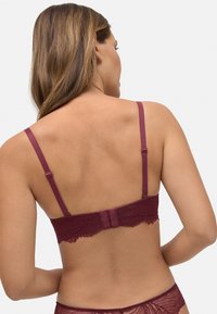 Sostén de encaje burdeos con tirantes ajustables, detalles de borde festoneado y cierre de gancho y ojo en la parte posterior. Textura suave en general.