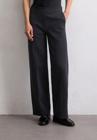 Schwarze weit geschnittene Hose aus weichem Stoff mit glatter Oberflächenstruktur. Mit hoher Taille und Seitentaschen, kombiniert mit polierten schwarzen Schuhen.