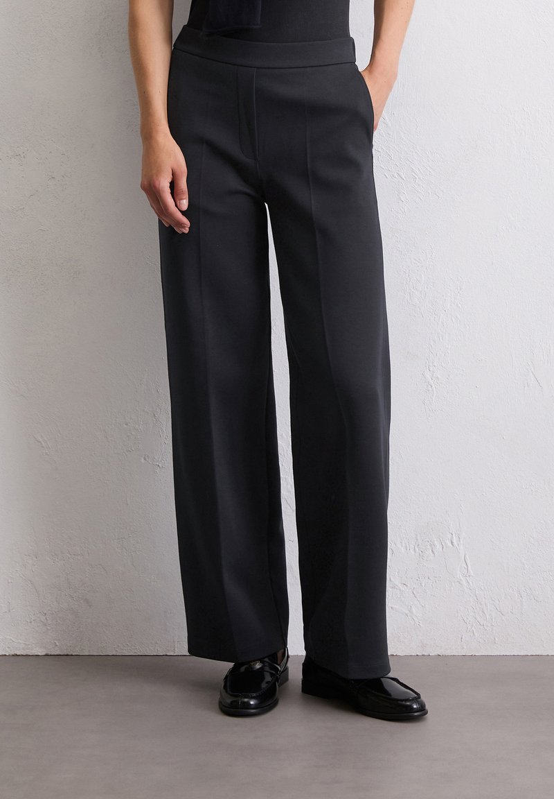 Schwarze weit geschnittene Hose aus weichem Stoff mit glatter Oberflächenstruktur. Mit hoher Taille und Seitentaschen, kombiniert mit polierten schwarzen Schuhen.