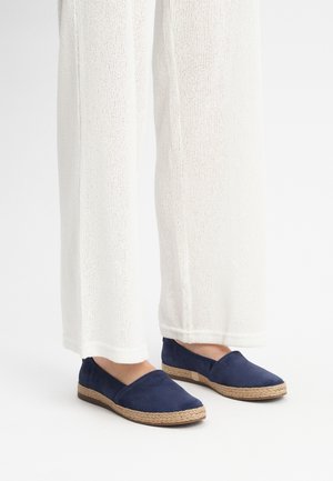Navy espadrilles with a soft suede upper and woven jute sole. Paired with loose, light-colored white trousers.