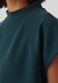 Top teal a maniche corte con collo alto, caratterizzato da una texture liscia e dettagli sottili di cuciture, che enfatizzano una vestibilità rilassata intorno al collo.