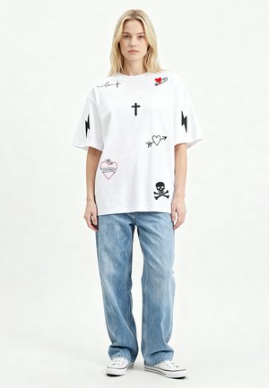 Blonde vrouw staat, draagt een wit oversized T-shirt met zwarte en rode tatoeage-achtige ontwerpen en losse blauwe jeans met witte sneakers.
