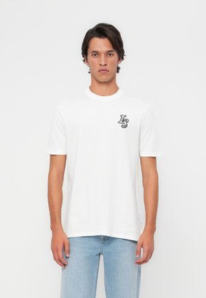 T-shirt en coton blanc avec des manches courtes, col rond, arborant un petit motif graphique noir sur la poitrine gauche. Le mannequin porte un jean bleu clair.