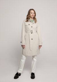 Beige trenchcoat met een ceintuur in de taille en knoopdetails, gecombineerd met witte broek en zwarte schoenen. Soepele textuur, klassiek ontwerp.