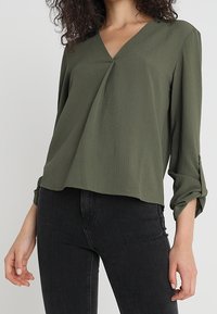Blusa verde oliva, a vestibilità ampia, con scollatura a V, maniche lunghe e polsini arrotolati. Tessuto testurizzato con pieghe sottili sul davanti.