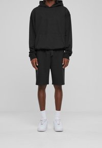 Sudadera negra con capucha y bolsillo frontal, combinada con pantalones cortos negros hasta la rodilla. Llevado con zapatillas blancas y calcetines blancos de caña corta.