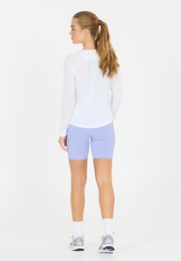 Vit långärmad träningströja kombinerad med ljuslila cykelshorts. Modellen står med ryggen mot kameran och visar upp en passande silhuett och en avslappnad stil.