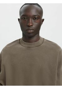 Brūna lielizmēra džemperis ar ribotu apaļu kakla izgriezumu, kam ir gluda tekstūra un smalka spīduma. Krūte ir vaļīga un minimālistiska.