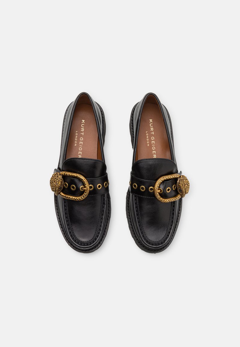 Une paire de mocassins en cuir noir avec des boucles décoratives en or et des œillets, des coutures gaufrées, et des semelles intérieures marron portant l'étiquette Kurt Geiger London.