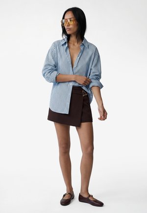 Kvinde iført en oversized blåstribet skjorte, sort asymmetrisk nederdel med spænde, brune flade sko og ravfarvede briller, stående med armene over kors.
