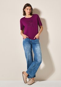 T-shirt violet à manches courtes avec impression noire, associé à un jean taille haute bleu clair à jambes larges et des chaussures basses beige. Fini textile lisse.