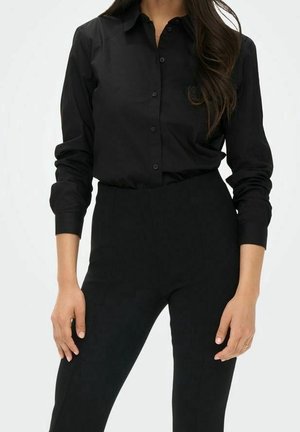 Femme portant une chemise noire cintrée à boutons avec manches longues et un pantalon noir taille haute, debout les mains relâchées le long du corps.