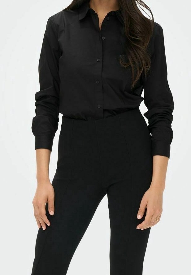 Femme portant une chemise noire cintrée à boutons avec manches longues et un pantalon noir taille haute, debout les mains relâchées le long du corps.
