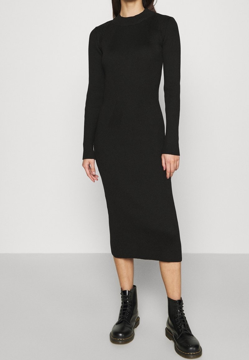 Robe longue à manches longues en maille côtelée noire avec un col montant, une silhouette ajustée et une longueur mi-mollet, associée à des bottes noires à lacets.