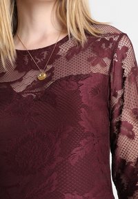 Burgundské krajkové tričko se květinovými vzory, průhlednými rukávy a kulatým výstřihem, doplněné jednoduchým zlatým náhrdelníkem.