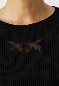 Černý žebrovaný pletený top s průhledným logem uprostřed. Výstřih je široký a kulatý, zvýrazněný vertikálními texturovanými vzory.