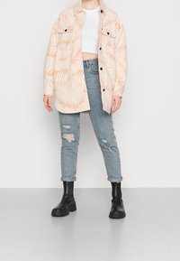 Camicia oversize color crema chiaro con motivo a foglie pesche, abbinata a jeans chiari strappati e stivali neri alla caviglia.