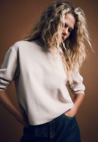 Blondynka z długimi kręconymi włosami, ubrana w biały sweter i niebieskie dżinsy, z dłońmi w kieszeniach, patrzy w dół na tle brązowym.